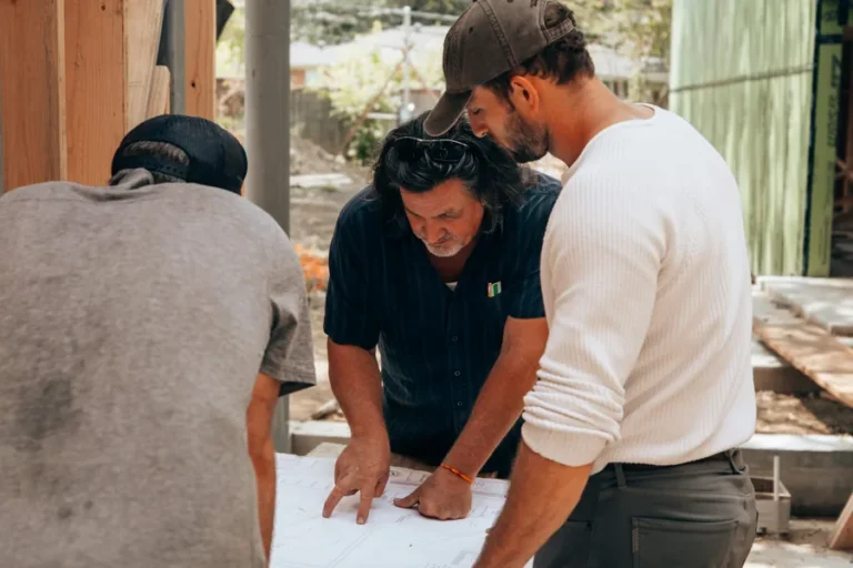 Three contractors leaning over a set of building plans together on a timber-frame construction site