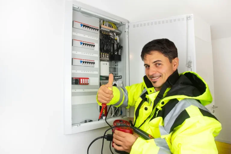 Electrician in a hi-vis jacket giving a thumbs up beside a newly wired consumer unit, holding a multimeter