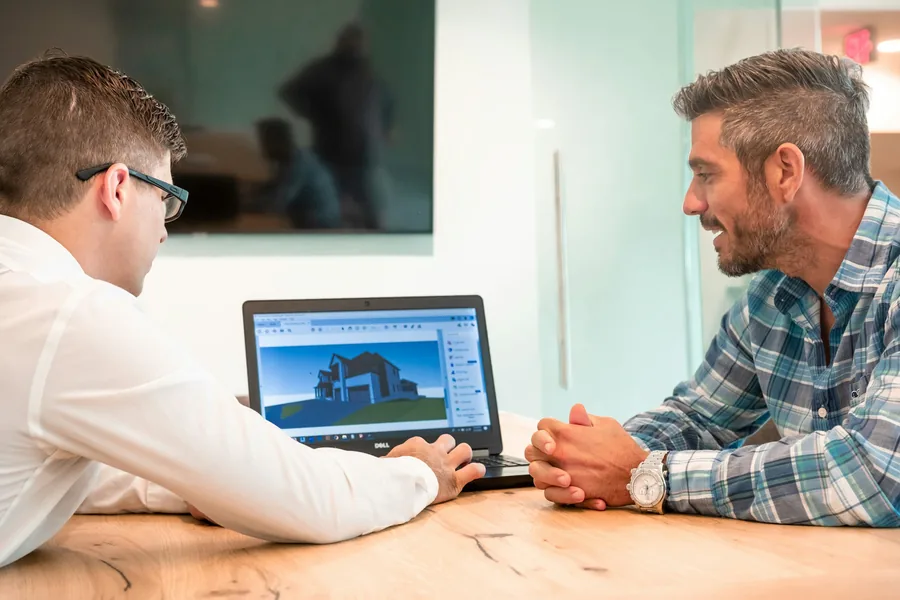 Adviser showing a smiling tradesman a house design on a laptop during a meeting at a wooden table