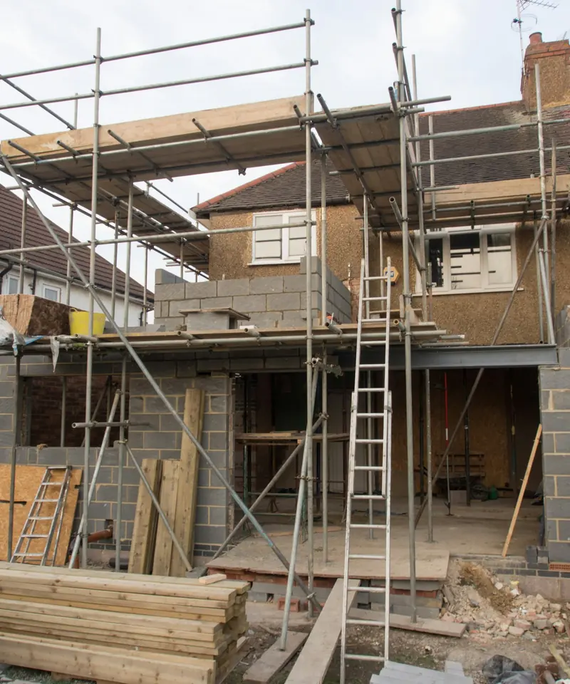 Two-storey rear extension under construction on a UK semi-detached house, with scaffolding, blockwork and stacked timber in the garden